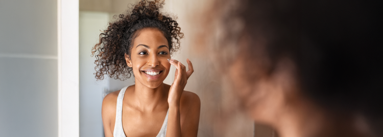 Blog of Transform Your Skin: Science-Backed Supplements for a Radiant Complexion which have image A woman with curly hair pulled up in a high bun smiles as she applies skincare cream to her face while looking in the mirror.