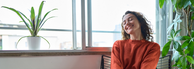 Blog of Magnesium Glycinate vs. Citrate: What’s the Difference? which have image A woman in an orange sweater sits in a bright room near a window, smiling with her eyes closed as she basks in the natural sunlight. 