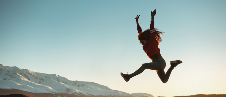 Blog of The Best Vitamins and Supplements to Boost Energy (Naturally) which have image A young woman jumps energetically in the air against a stunning backdrop of snow-capped mountains during sunset, symbolizing freedom, adventure, and happiness.
